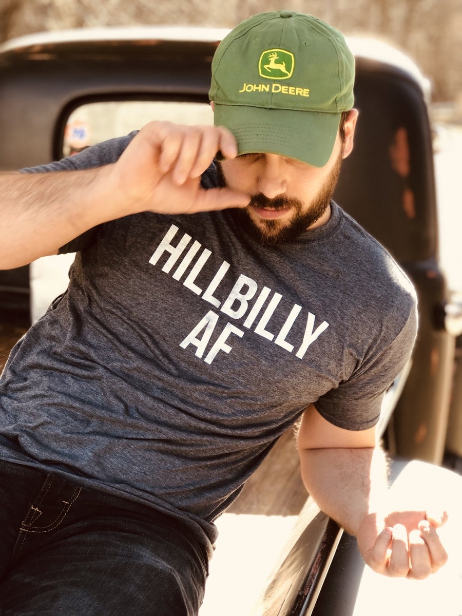 Man sitting on the back of a truck wearing a green hat and a grey Hill and Holler Shirt.
